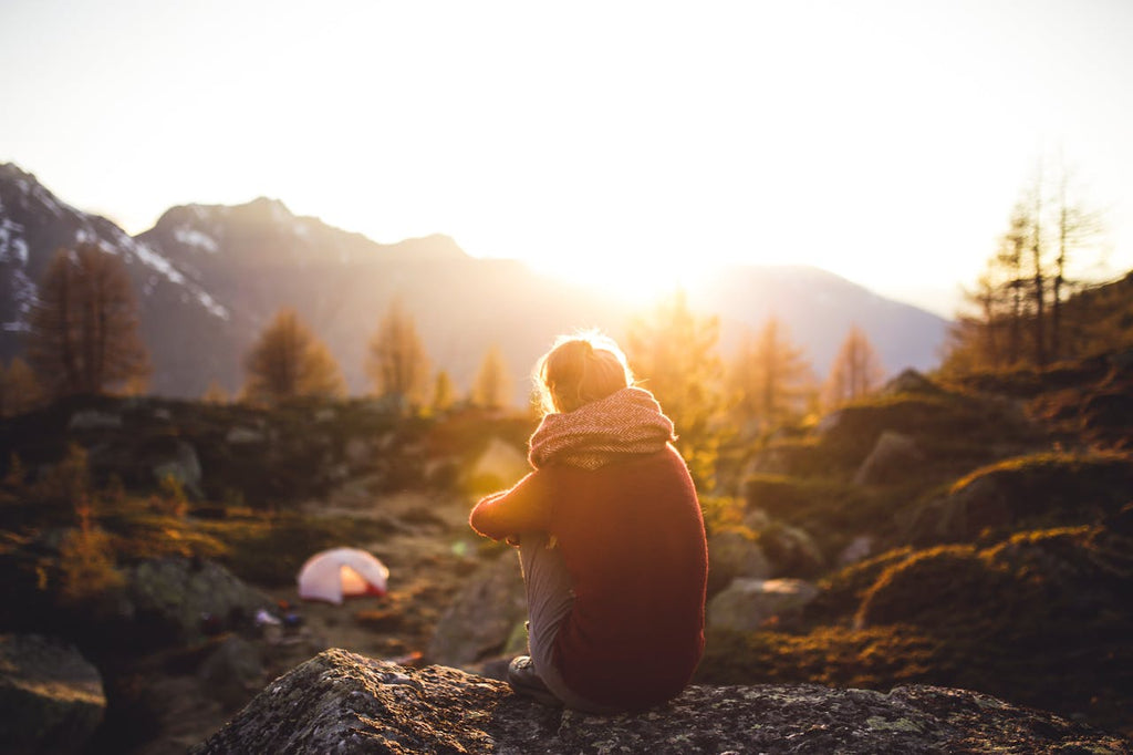 Person enjoying a peaceful moment in nature at sunset while sitting on a rock by a campsite.