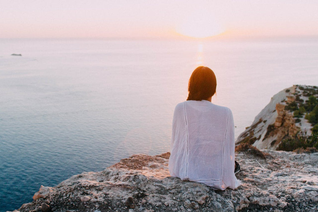 Woman sitting on a rocky ledge overlooking the sea at sunset, embodying tranquility and mindfulness.